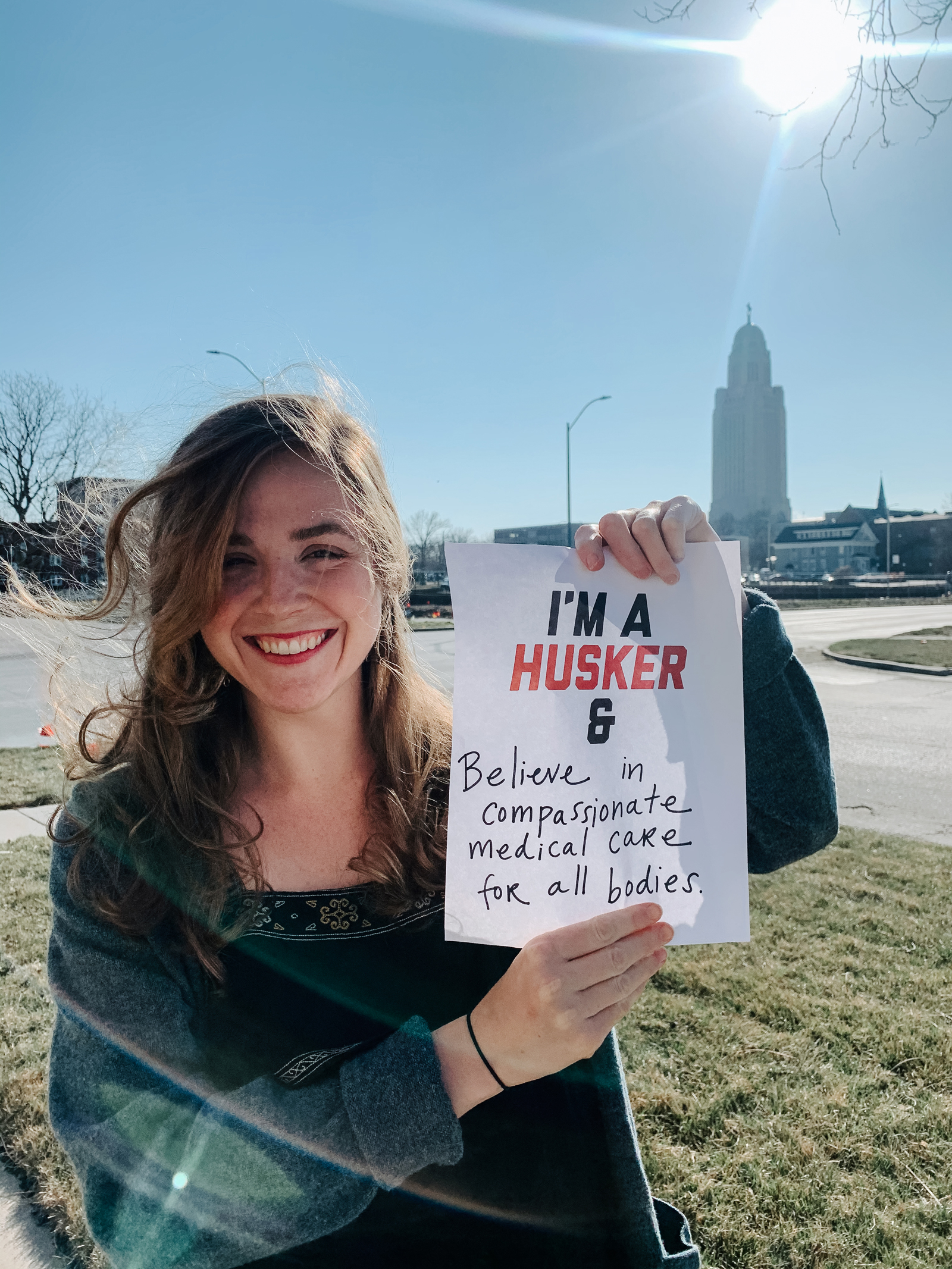 Jacqueline smiles for the camera with the Capitol in the background. She holds a sign that says “I’m a Husker & I believe in compassionate medical care for all bodies”