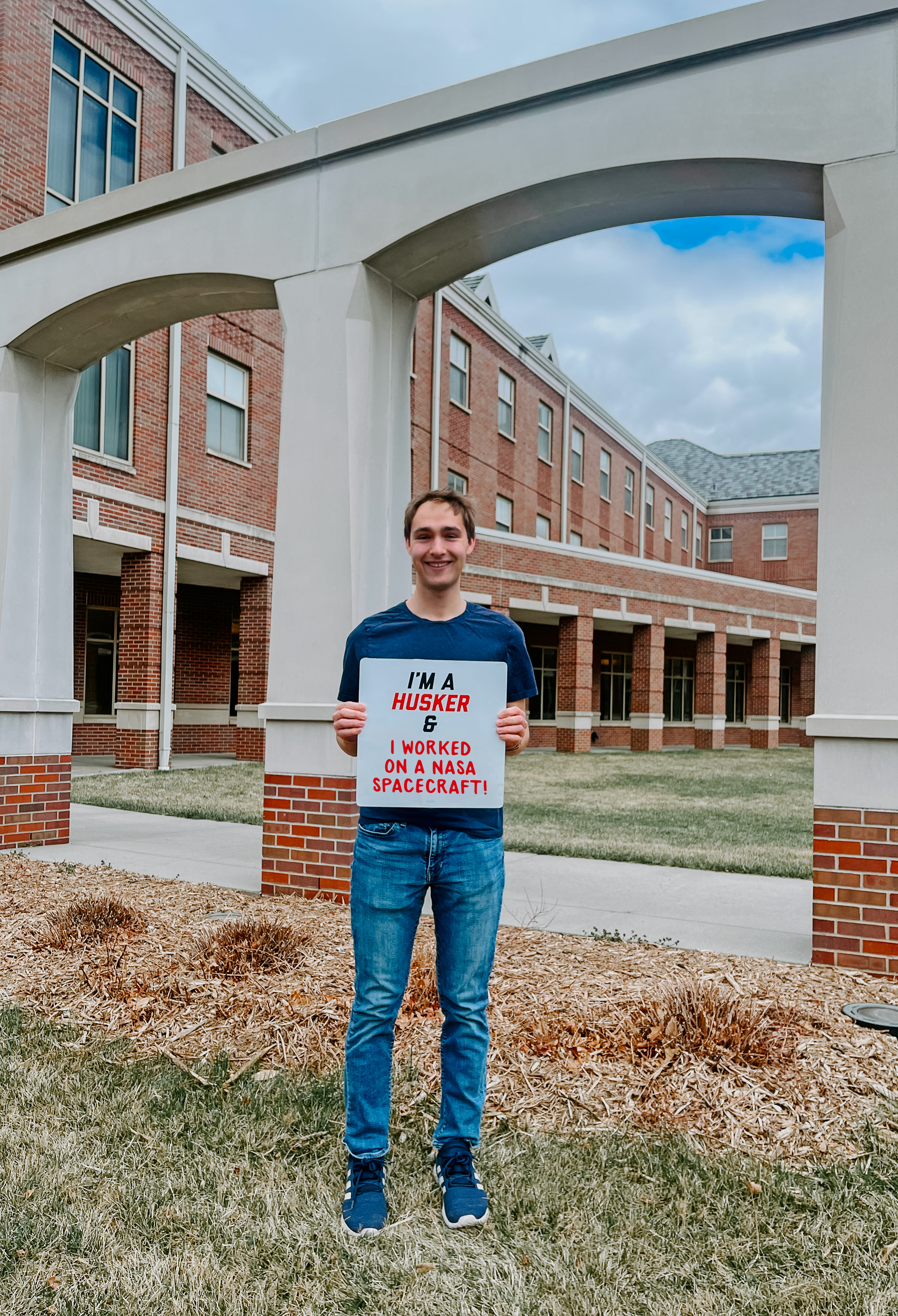 Ben stands outside the Raikes building and holds a sign reading "I'm a Husker and I've worked on a NASA spacecraft"