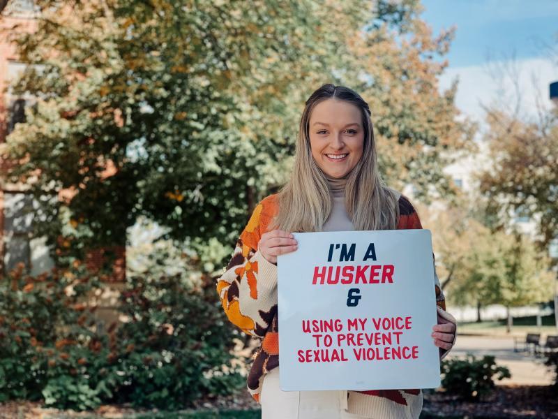 Em smiles for a photo outside holding a board that says "I'm a Husker & using my voice to prevent sexual violence"