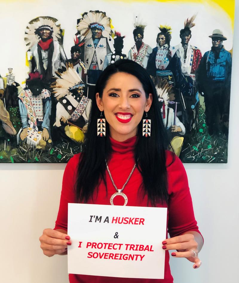 Katie smiles for a photo holding a sign that says "I'm a Husker & I protect tribal sovereignty"