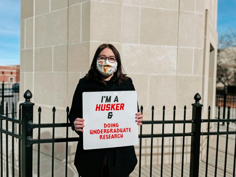 Delainie holds a sign that says "I'm a Husker & doing undergraduate research"