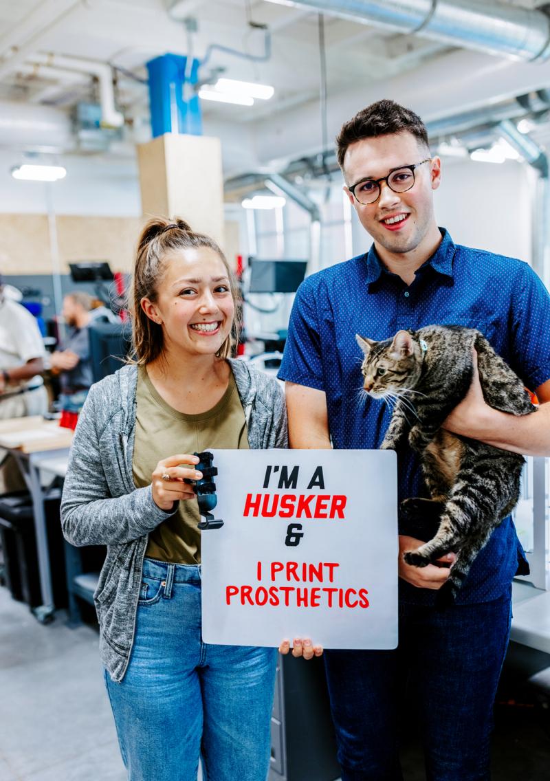 Abby and Harrison pose for a photo with Olive the cat in the Maker’s Studio. They hold a sign that says “I’m a Husker and I print prosthetics”