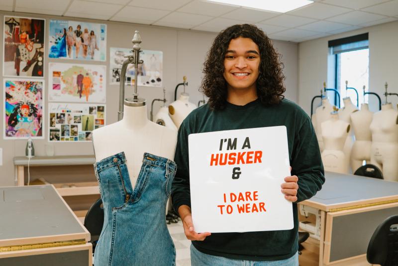 Delano smiles for a photo with his denim dress and a sign that says “I’m a Husker & I dare to wear”