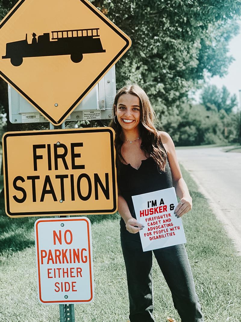Laura poses for a photo with a sign that says “I’m a Husker & firefighter cadet & advocating for people with disabilities”