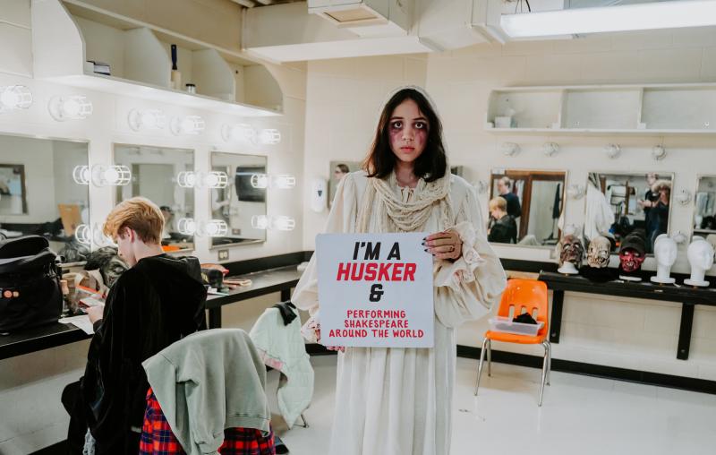 Aurora poses for a photo in the dressing room in their ShakesFEAR costume. She holds a sign that says “I’m a Husker & performing Shakespeare around the world”