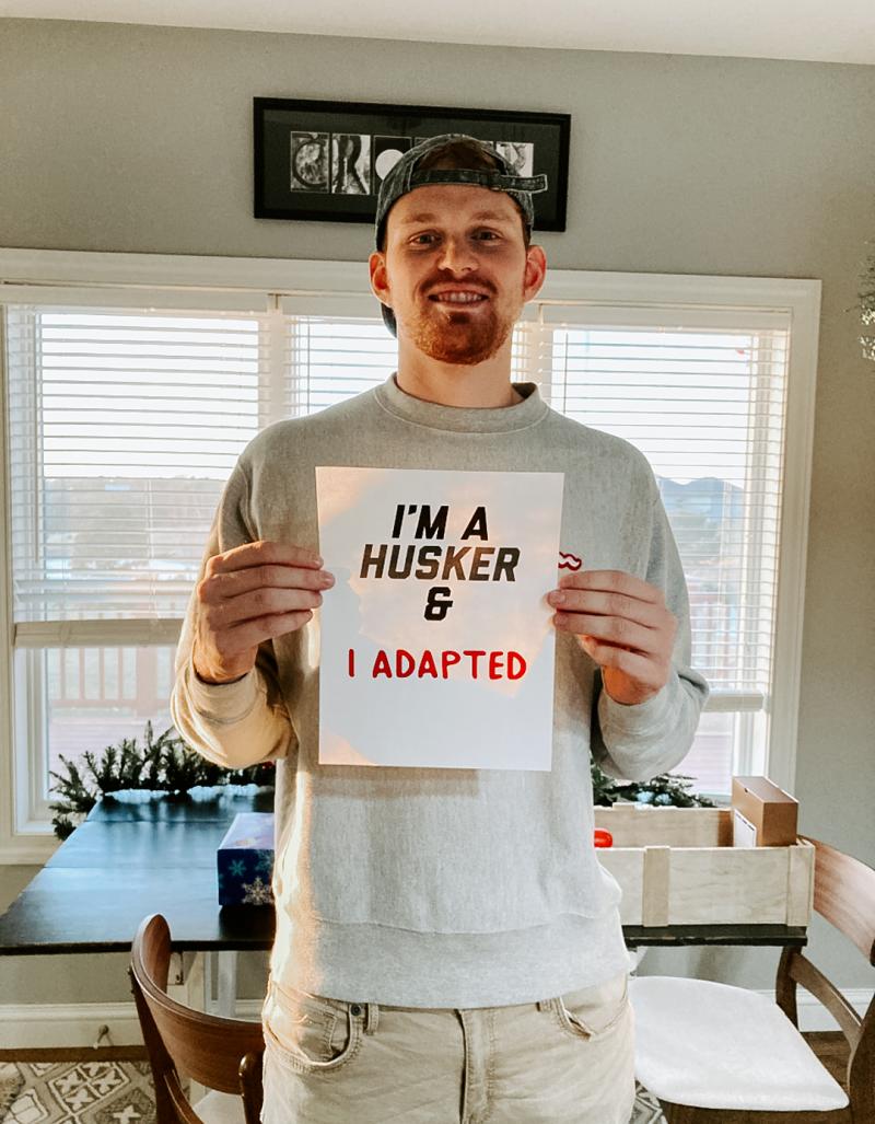 Alec smiles for a photo at home, holding a sign that says "I'm a Husker & I adapted"