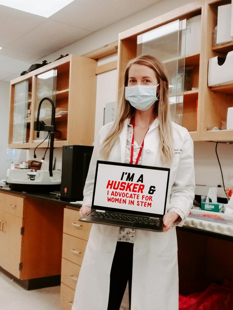 Courtney stands in a lab on campus holding a computer that says “I’m a Husker & I advocate for women in STEM”