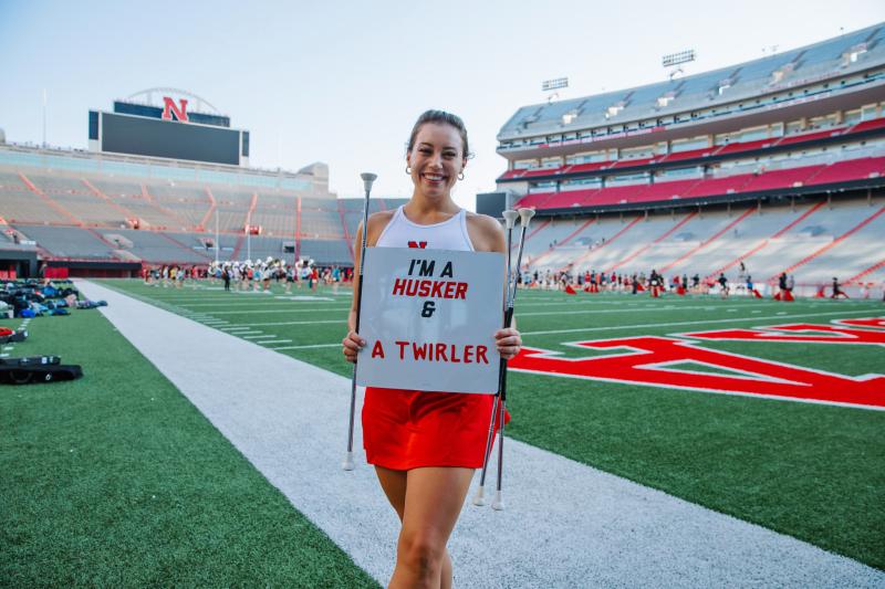 Steffany smiles for a photo on the field with a sign that says "I'm a Husker & a twirler"