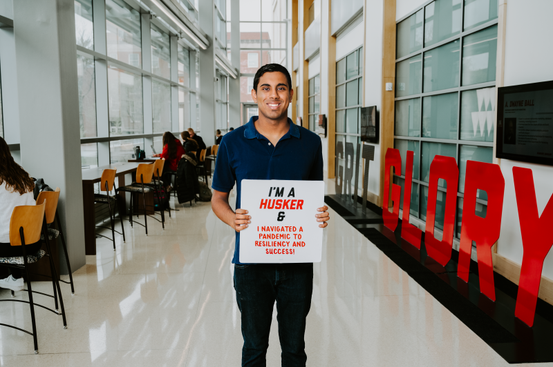 Ananth stands in a hall with all windows at UNL College of Business. He holds a sign reading "I'm a Husker and I navigated a pandemic to resiliency and success!"