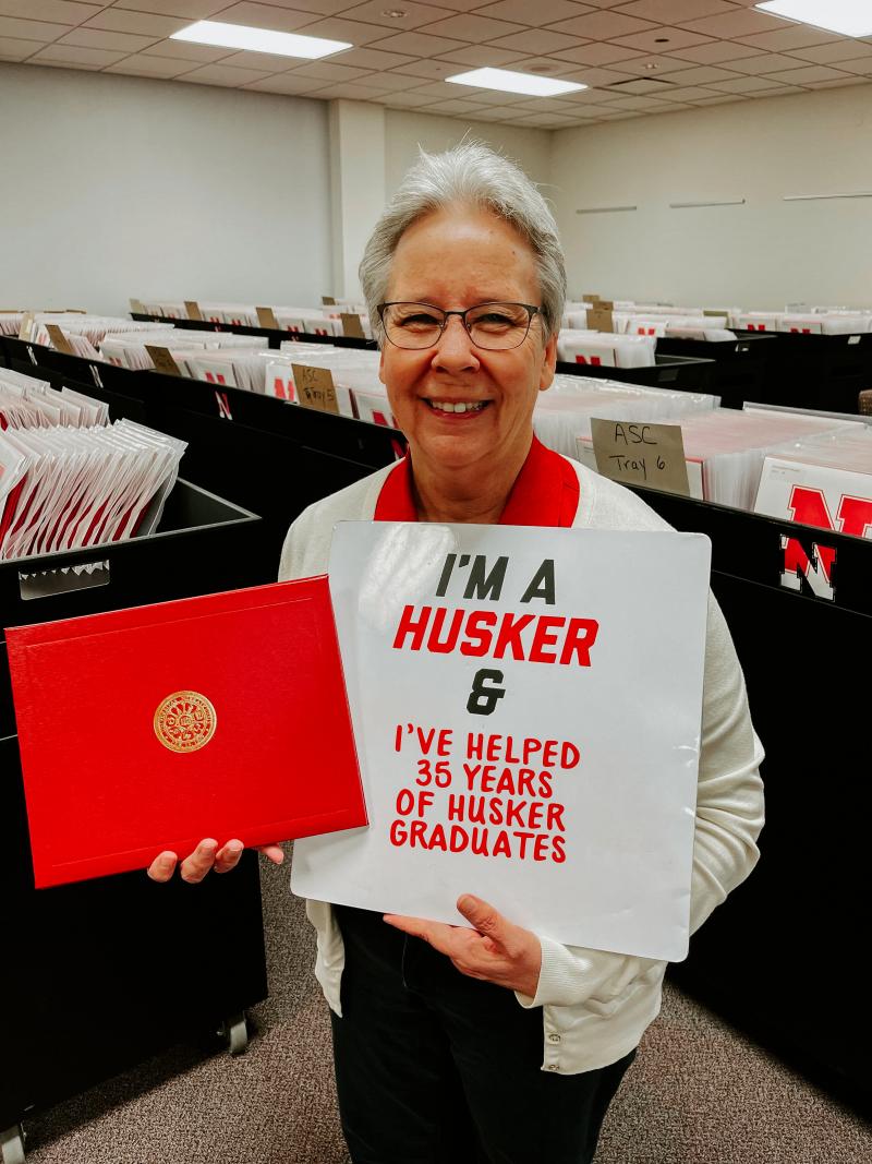 Gail stands in a room with hundreds of diplomas. She holds a sign reading, "I'm a Husker and I've helped 35 years of Husker graduates"