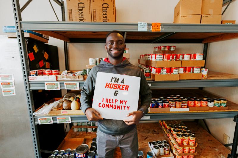 Gabin smiles for a photo in the pantry, holding a sign that reads "I'm a Husker & I help my community."