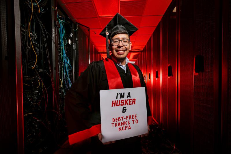 Bryan smiles for a photo in his grad regalia, holding a sign that says "I'm a Husker & debt-free thanks to NCPA”