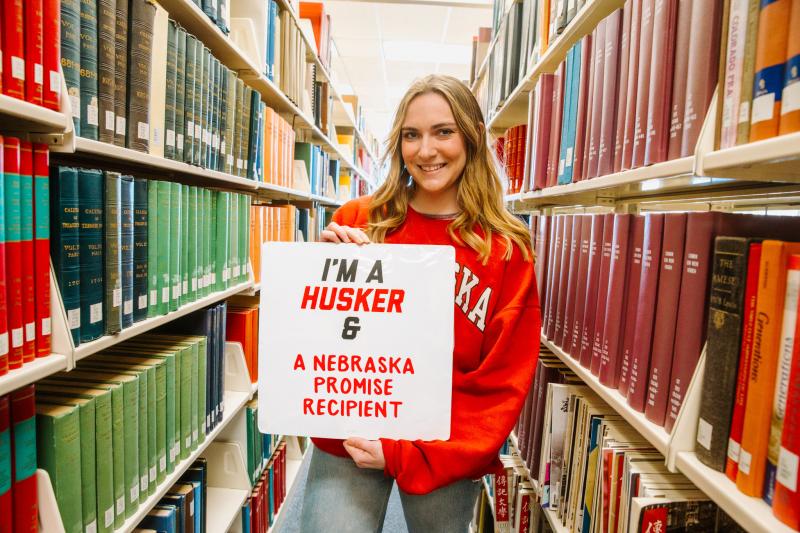 Nonie smiles for a photo between the stacks in the library holding a sign that reads "I'm a Husker & a Nebraska Promise Recipient"