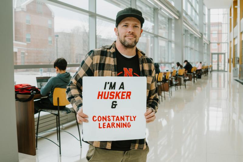 Mitch smiles for a photo holding a board that reads "I'm a Husker & constantly learning"