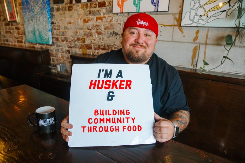 Nick smiles for a photo inside Muchachos with a sign that says "I'm a Husker & building community through food"