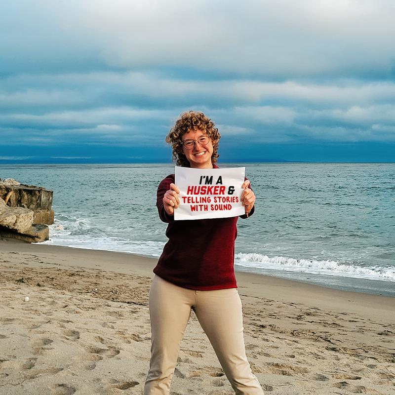 Alijah smiles for a photo on a beach during her internship. She holds a sign that says "I'm a Husker & telling stories with sound"