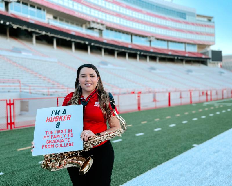 Aracely stands on the football field at Memorial Stadium and holds a sign reading "I'm a Husker and the first in my family to graduate from college"