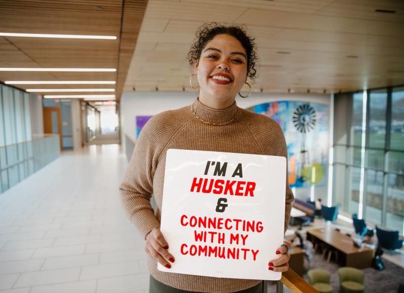 Azcia smiles for a photo with a board that reads "I'm a Husker & Connecting with my Community"
