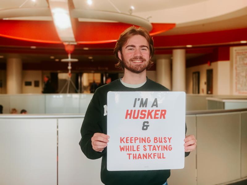 Brendan smiles for a photo holding a sign that says "I'm a Husker & keeping busy while staying thankful"