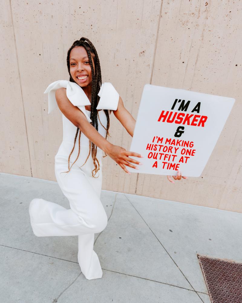 Cherish wears an all white jumpsuit and holds a sign reading, "I'm a Husker and I'm changing the world one outfit at a time"