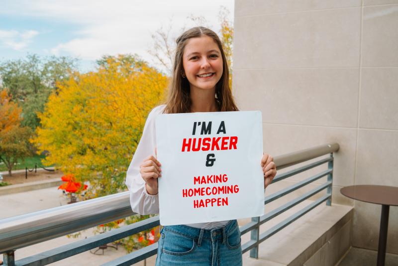 Chrissy smiles for a photo on the Union balcony with a board that reads "I'm a Husker & making homecoming happen"