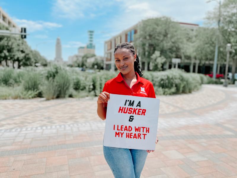 Clare stands in front of the Nebraska Capitol and surrounding landscaping. She holds a sign reading "I'm a Husker and I lead with my heart"