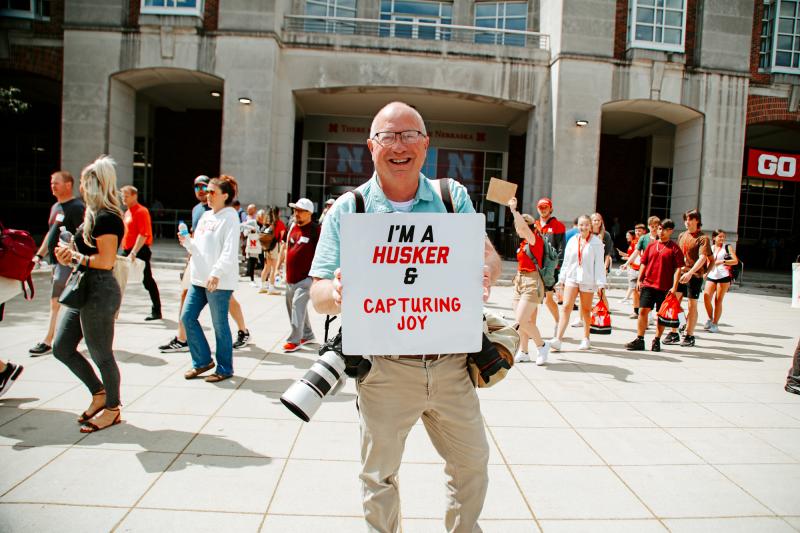 Craig poses for a photo outside the Union with incoming students walking behind him. He holds a sign that says "I'm a Husker & capturing joy"