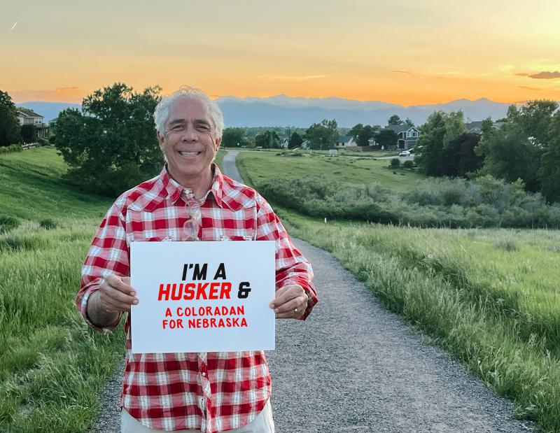 Dan smiles for a photo as the sun sets with mountains in the background. He holds a sign that reads "I'm a Husker & a Coloradan for Nebraska"
