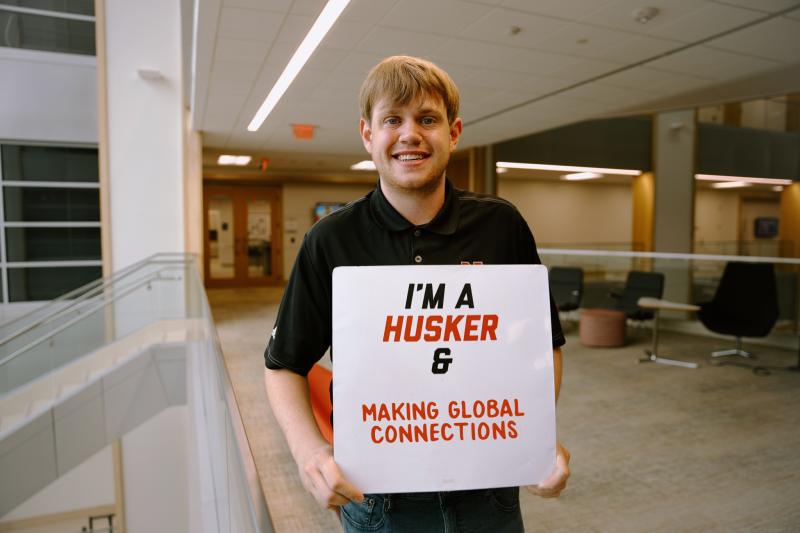 Devan smiles for a photo in Hawks Hall with a board that reads “I’m a Husker & making global connections”
