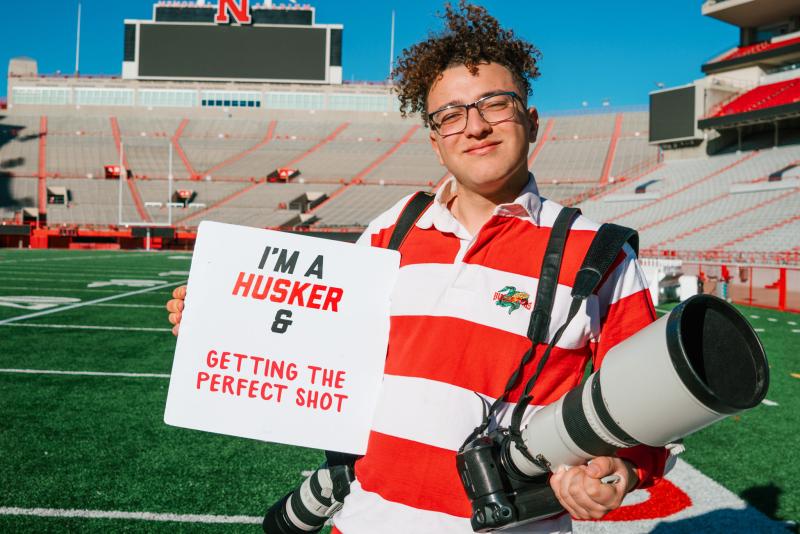 Dillon smiles for a photo holding his cameras and a sign that says "I'm a Husker & Getting the Perfect Shot"