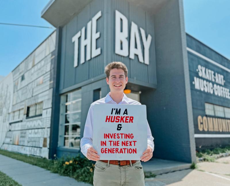 Ethan stands in front of The Bay and holds a sign reading "I'm a Husker and investing in the next generation"