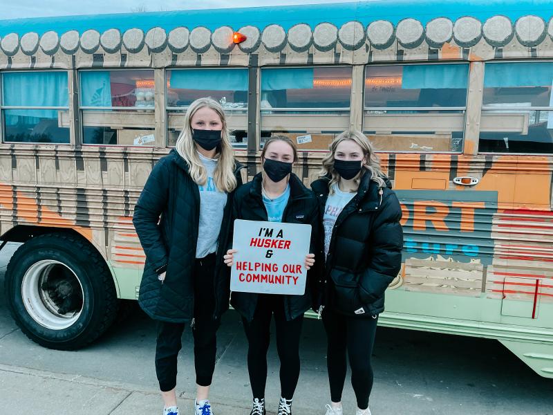 Maddie, Kaelyn and Emma pose for a photo outside the Food Fort bus holding a sign that reads "I'm a Husker & helping our community"