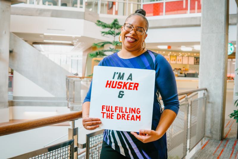 Genese smiles for a photo holding a sign that says "I'm a Husker & fulfilling the dream"