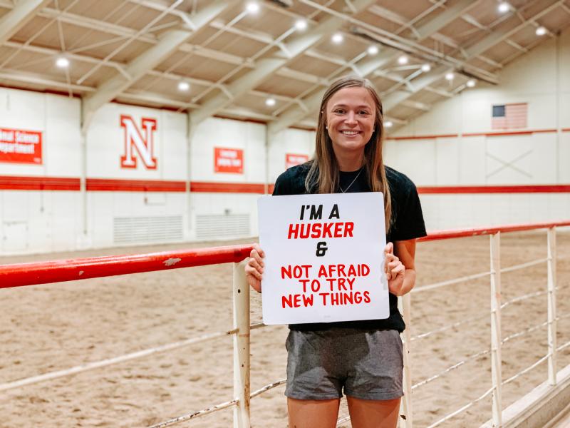 Hadley smiles for a photo in the equestrian arena holding a sign that says "I'm a Husker & not afraid to try new things"