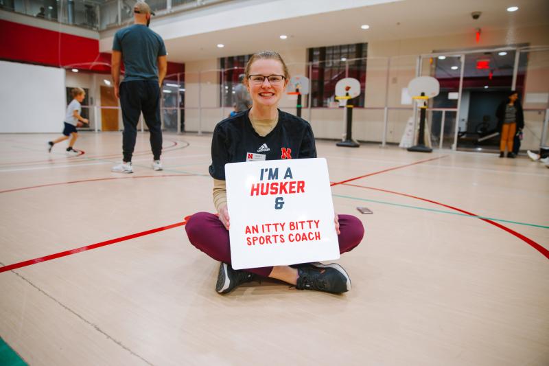 Hailey smiles for a photo in the gym holding a board that reads "I'm a Husker & an itty bitty sports coach"