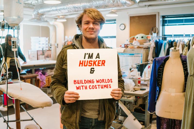 Hunter smiles for a photo amongst the costumes as he holds a sign that reads “I’m a Husker & building worlds with costumes”