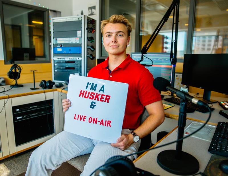 Jacob smiles for a photo in the KRNU studio as he holds a sign that reads “I’m a Husker & live on-air”
