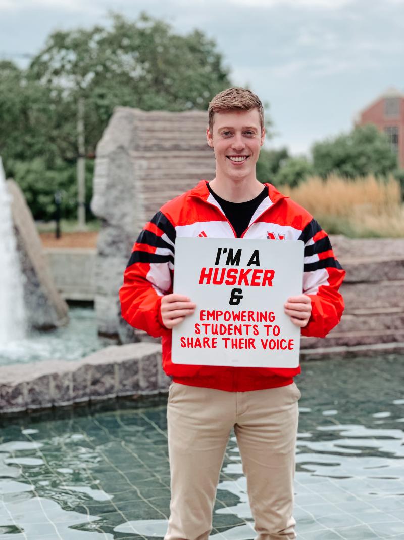 Jake smiles for a photo with a sign that says "I'm a Husker & empowering students to share their voice"