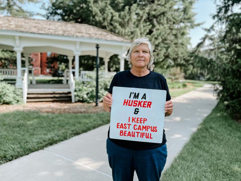 Jolene stands in front of Perin Porch at East Campus. She holds a sign reading, "I'm a Husker and I keep East Campus beautiful."