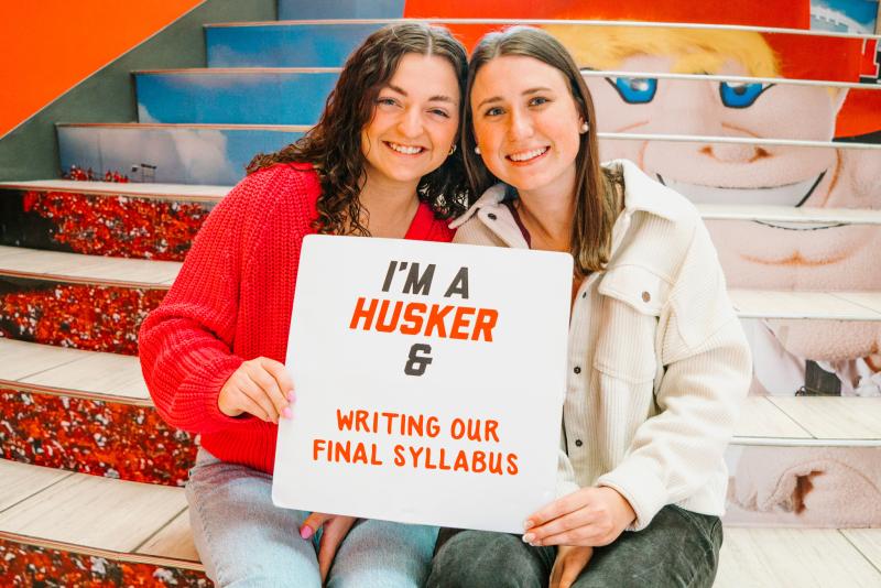 Katie and Sarah smiles for a photo on stairs inside the Nebraska Union. They hold a sign that reads "I'm a Husker & writing our final syllabus" 