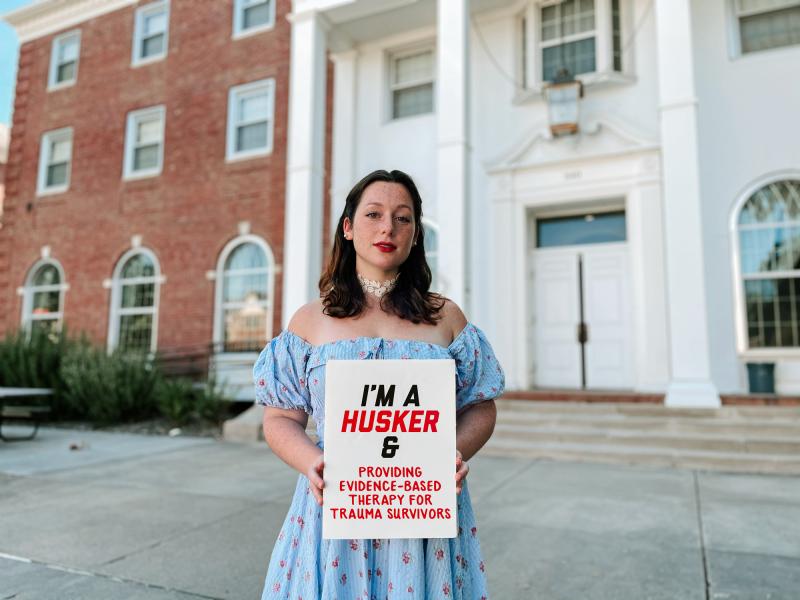 Katie takes a photo outside of the Neihardt Center with a board that says "I'm a Husker & providing evidence-based therapy for trauma survivors"