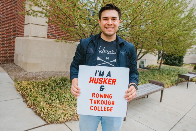 Kevin smiles for a photo with a sign that says "I'm a Husker & rowing through college"