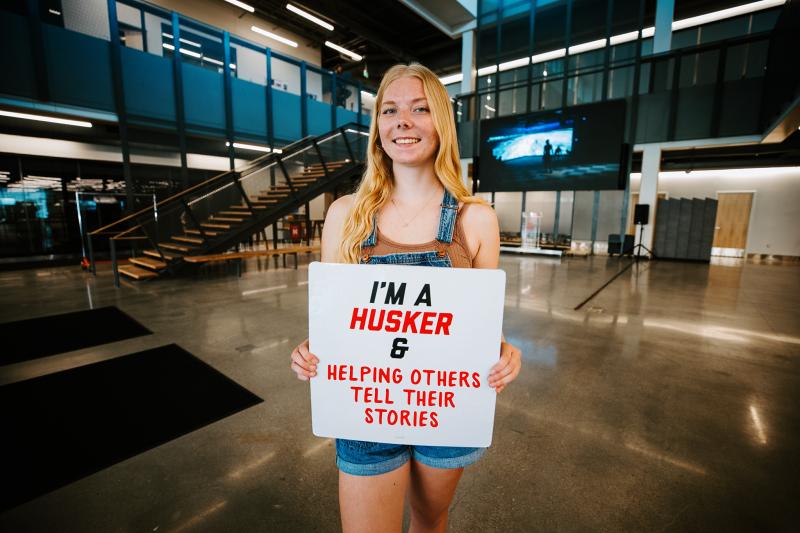 Kylie smiles for a photo in the Carson Center with a sign that reads "I'm a Husker & helping others tell their stories"
