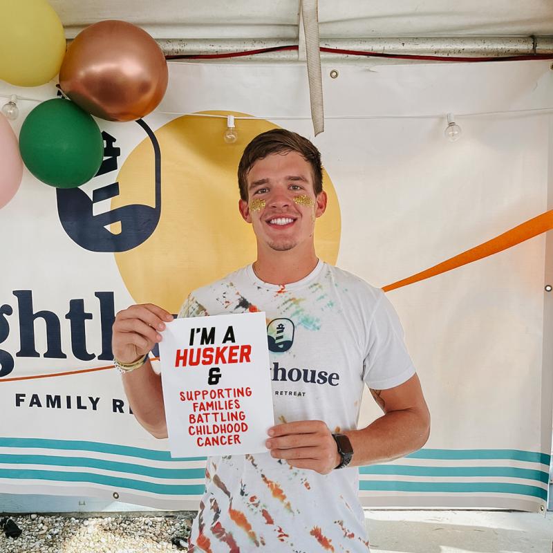 Luke stands in front of balloons and a billboard reading "Lighthouse Family Retreat" . He holds a sign reading "I'm a Husker & supporting famlies battling childhood cancer"
