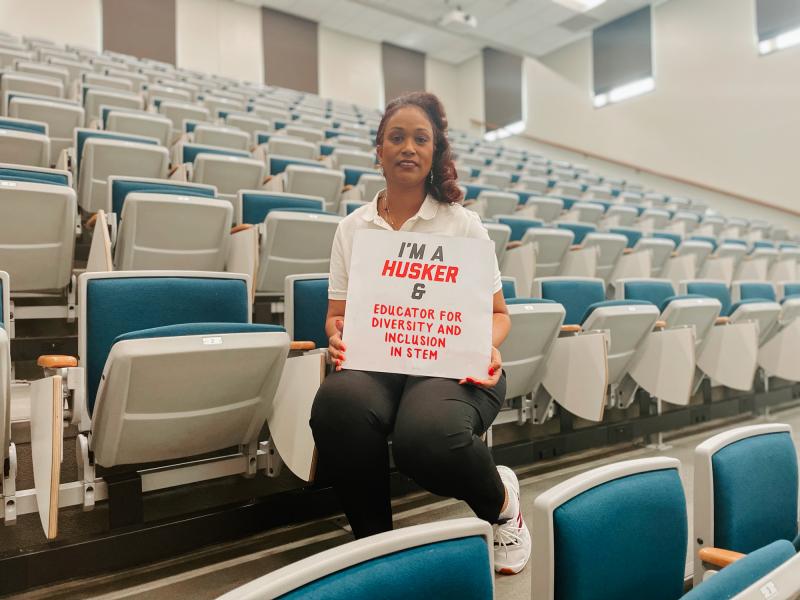 Marianna sits in a classroom holding a sign that says "I'm a Husker & educator for diversity and inclusion in STEM"
