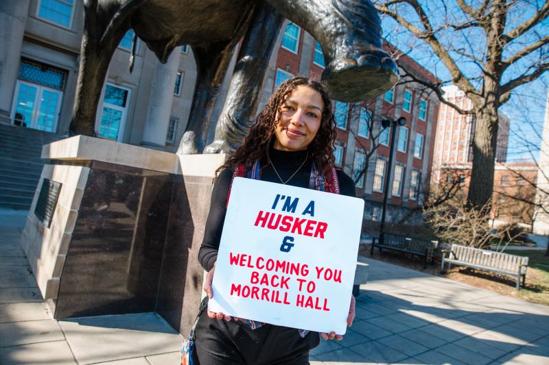 Maya smiles for a photo outside Morrill Hall with a sign that says "I'm a Husker & Welcoming You Back to Morrill Hall"