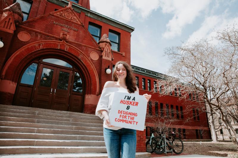 Meagan smiles for a photo outside Arch Hall holding a sign that says “I’m a Husker & designing for the community”