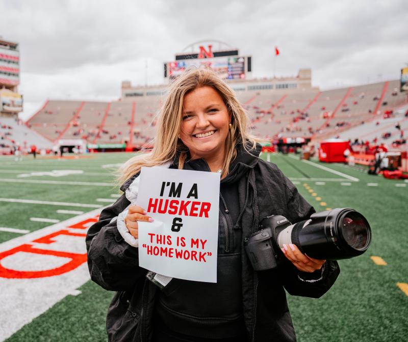 Meg smiles on the field before the spring game holding a camera in one hand and sign in the other. It reads: “I’m a Husker & this is my homework”