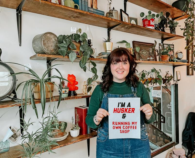 Megan stands in front of a wall decorated with plants and antique items. She holds up a board reading "i'm a Husker & Running my Own Coffee Shop." 