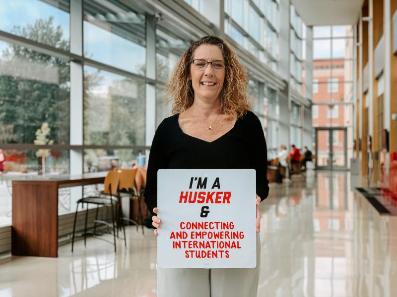 Mikki smiles for a photo in the College of Business holding a sign that reads “I’m a Husker & connecting and empowering international students”
